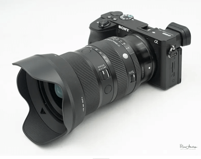 A black Sony Alpha mirrorless camera with a large zoom lens and lens hood attached, placed on a white surface.